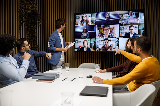 Candid shot of group of businessmen sitting and standing around a conference table in a board room, having a video conference via flat screen TV with their colleagues.