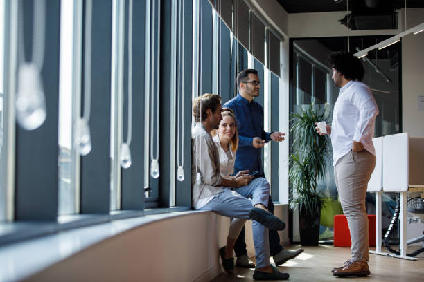 Full length shot of four business colleagues hanging out by the windows in the modern, open plan office space, chatting and relaxing during coffee break.