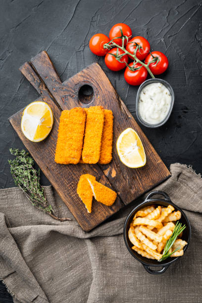 Fried fish finger stick or french fries fish with sauce, on wooden cutting board, on black background, top view flat lay Fried fish finger stick or french fries fish with sauce set, on wooden cutting board, on black background, top view flat lay goldfish snack stock pictures, royalty-free photos & images