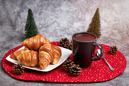 Cup of hot chocolate with a plate with croissant on a red Christmas tablecloth and some Christmas fir trees. Christmas Concept