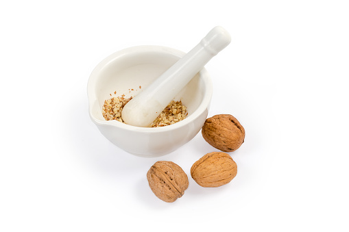 Crushed kernels of walnuts in the white porcelain kitchen mortar with pestle and whole unshelled nuts on a white background
