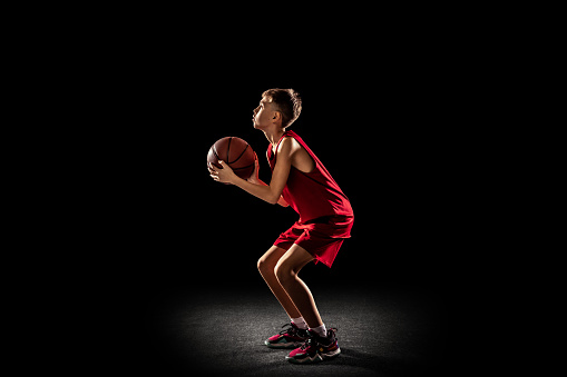 Hight level of concentration. Portrait of teen boy, playing, training basketball isolated on black background. Throwing ball. Concept of motivation, sport, action, movement, health. Copy space for ad