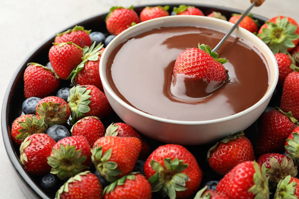 istock Fondue fork with strawberry in bowl of melted chocolate surrounded by different berries on light table, closeup 1356152949