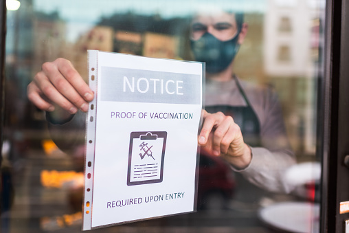 Young man hanging information sign “proof of vaccination required” on the entrance door of a restaurant.