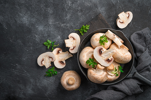 Cooking champignons, raw chopped and whole champignons in a frying pan on a dark background. Top view, copy space.