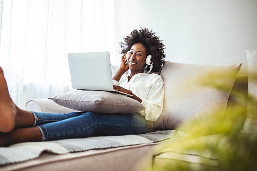 Beautiful young African woman in casual clothing enjoying music and smiling while resting at home. Young woman with headphones using laptop at home