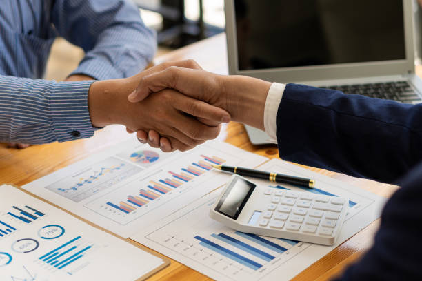 istock businesspeople shaking hands while standing near the office window after doing modern office cooperation agreement and financial paper graph on the table 1356162170