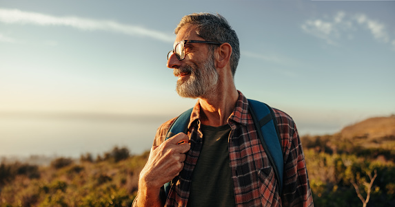 Enjoying the hilltop views. Happy mature hiker looking away with a smile on his face while standing on top of a hill with a backpack. Adventurous backpacker enjoying a hike at sunset.