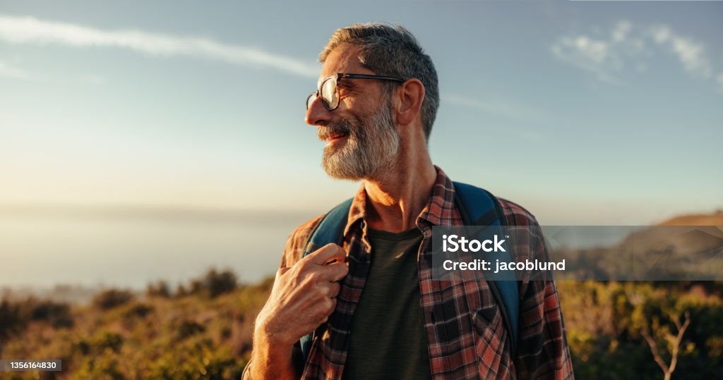 Enjoying the hilltop views Enjoying the hilltop views. Happy mature hiker looking away with a smile on his face while standing on top of a hill with a backpack. Adventurous backpacker enjoying a hike at sunset. Senior Adult Stock Photo