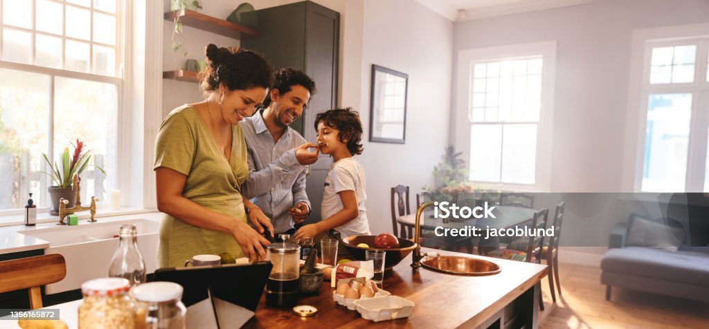 Playful dad feeding his son a slice of bread in the kitchen Playful dad feeding his son a slice of bread while his wife prepares breakfast. Family of three having fun together in the kitchen. Mom and dad spending quality time with their son. Family Stock Photo