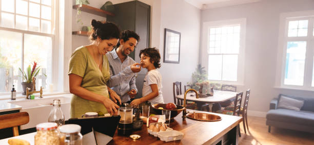 Playful dad feeding his son a slice of bread in the kitchen Playful dad feeding his son a slice of bread while his wife prepares breakfast. Family of three having fun together in the kitchen. Mom and dad spending quality time with their son. home stock pictures, royalty-free photos & images