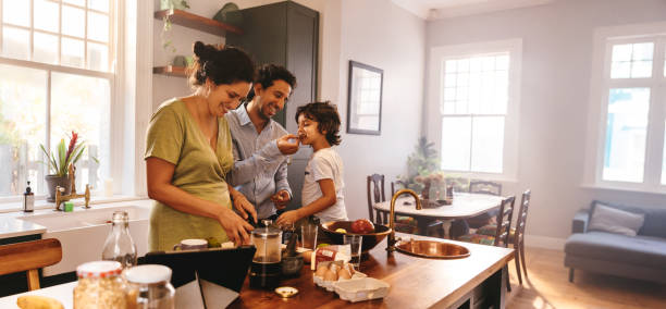 Playful dad feeding his son a slice of bread in the kitchen Playful dad feeding his son a slice of bread while his wife prepares breakfast. Family of three having fun together in the kitchen. Mom and dad spending quality time with their son. family get together stock pictures, royalty-free photos & images