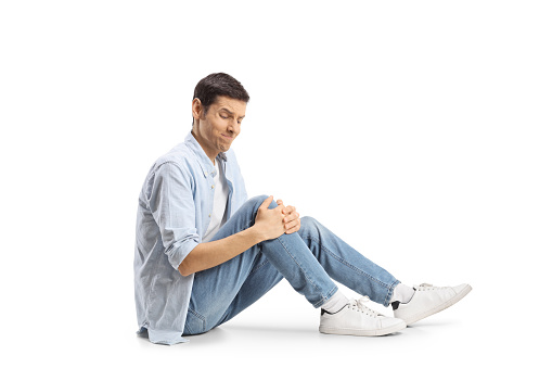 Young man sitting on the floor and holding his knee in pain isolated on white background