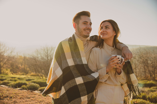 Copy space shot of affectionate young couple, walking embraced, under a cozy, warm blanket, in winter sunshine in beautiful scenic nature. Young woman is keeping her hands warm with a cup of tea and smiling at her boyfriend.