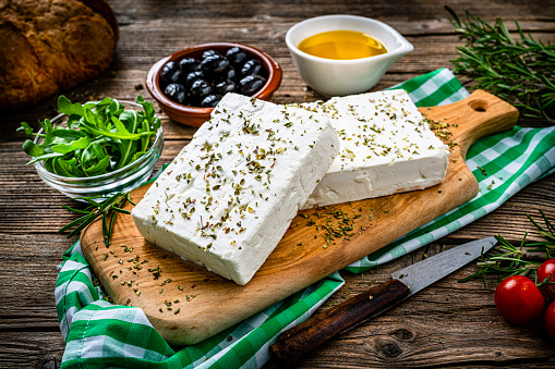 Regional food backgrounds: close up view of two blocks of Greek feta cheese on a cutting board shot on rustic wooden table. Olives, cherry tomatoes, olive oil, salt and pepper and herbs like rosemary, oregano, and arugula complete the composition. High resolution 42Mp studio digital capture taken with SONY A7rII and Zeiss Batis 40mm F2.0 CF lens