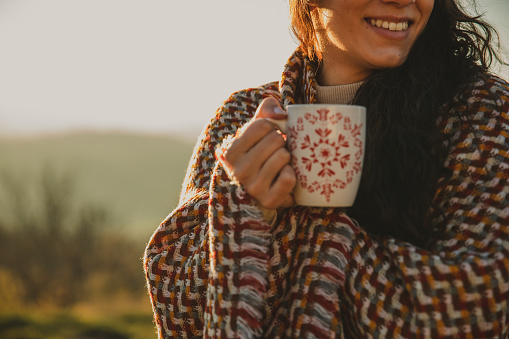 Cut out shot of smiling young woman wrapped in cozy blanket, holding a cup of tea, enjoying a sunny autumnal day outside.
