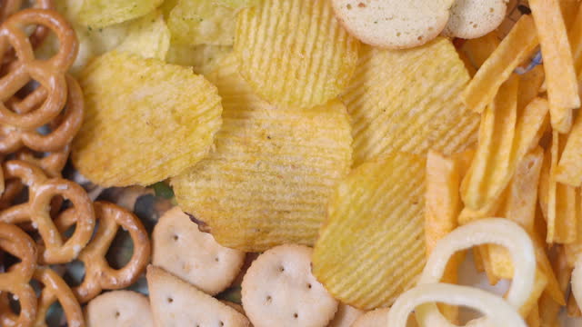 Many types of unhealthy salty snacks: chips, crackers, pretzels, onion rings. Top view. Rotation video