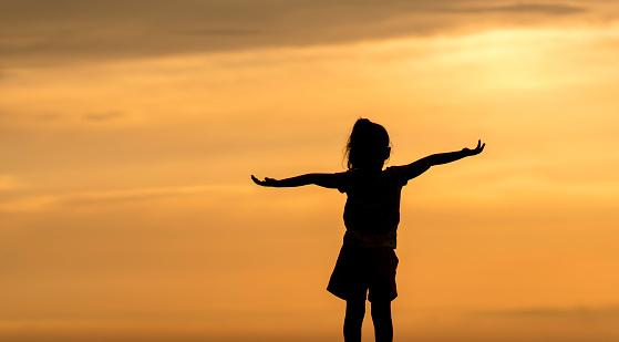 Silhouette of a little girl with hands raised in the sunset over the sea