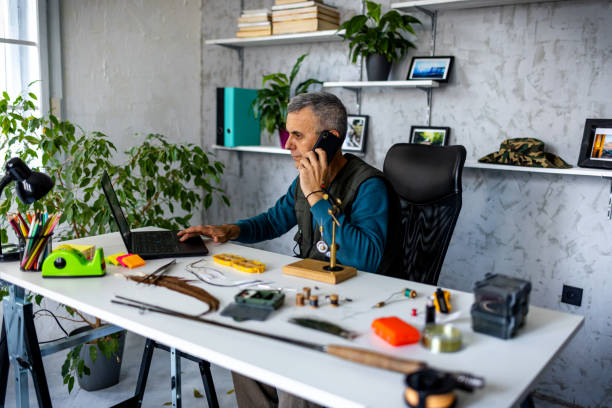 dono de pequeno negócio conversando com um cliente enquanto usa laptop em oficina - multi tasking craftsperson work tool men - fotografias e filmes do acervo