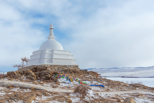 White Buddhist stupa and ritual trees with colorful ribbons on a cloudy winter day at the top of the sacred island of Ogoy. Lake Baikal, Siberia, Russia.