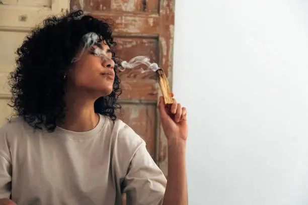 Young mixed race woman holding a smoking palo santo smudging herself and the room. Copy space. Spirituality and meditation concepts.