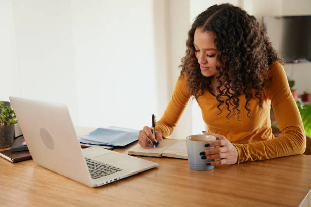istock Multi-cultural female making notes in notepad. Young professional remote working with laptop in modern apartment 1356177963