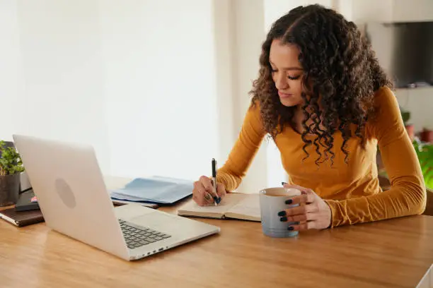 Multi-cultural female making notes in notepad. Young professional remote working with laptop in apartment.