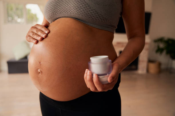 femmina caucasica in piedi in soggiorno che strofina la crema idratante sulla pancia incinta - ewan foto e immagini stock