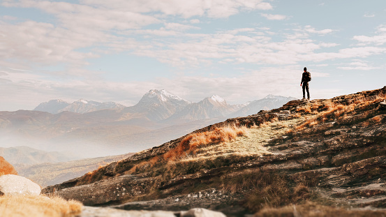 Adult man hiking solo and standing on top of a rock, looking at a majestic mountain range at the horizon. Warm light with beautiful sky. Total freedom and exploration.