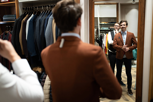 In the modern boutique, male customer trying new suit and observing himself, while skilled tailor and salesman assist to him