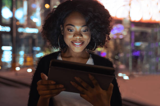 istock Woman looking at her tablet computer and smiling while standing at street at night time 1356194856