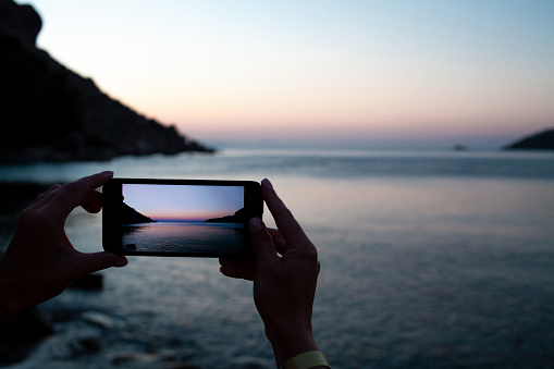 woman hands holding mobile phone at sunrise over sea bay. Taking photo of sunset over rocks in water by mobile phone. Selective focus on smartphone