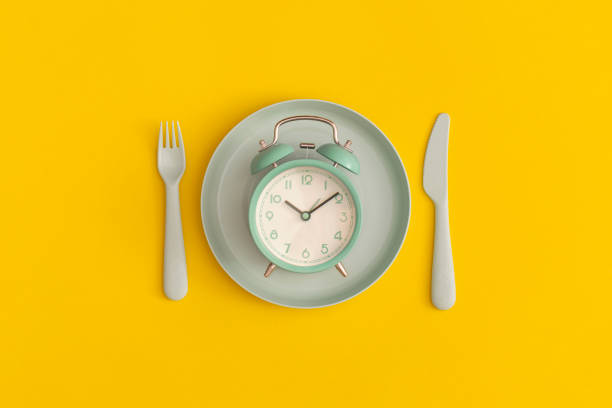 istock Green clock on the blue plate with fork and spoon on a yellow background. Top view. Flat lay. 1356196762
