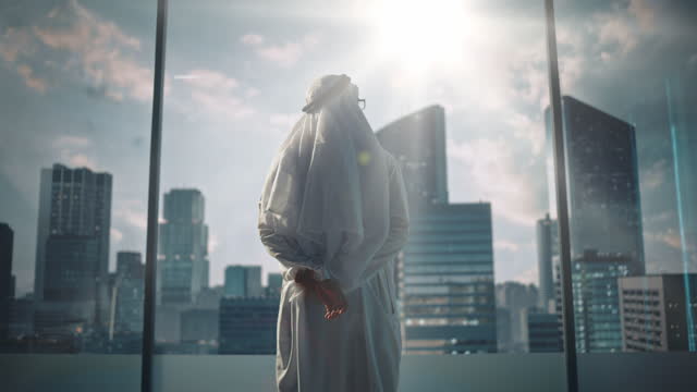 Successful Muslim Businessman in Traditional White Outfit Walking in His Modern Office Looking out of the Window on Big City with Skyscrapers. Successful Saudi, Emirati, Arab Businessman Concept.