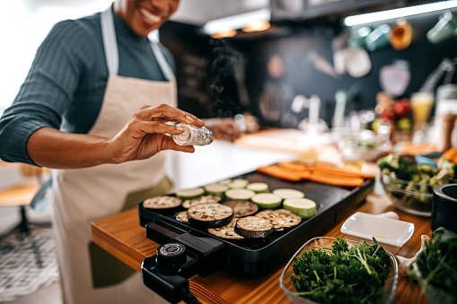 Happy woman pouring salt to eggplant slice while roasting on barbecue grill in kitchen