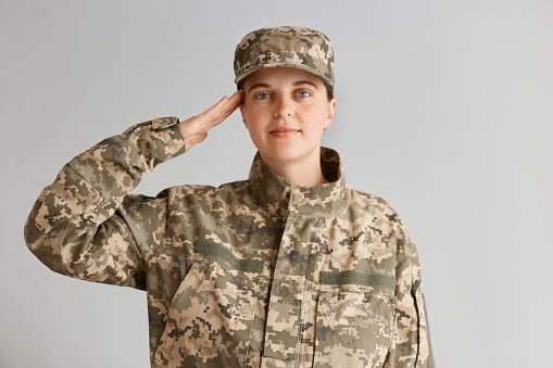 Indoor shot of positive woman soldier wearing camouflage uniform and cap, saluting with positive facial expression, standing against light backgrond, being glad to serve in army.