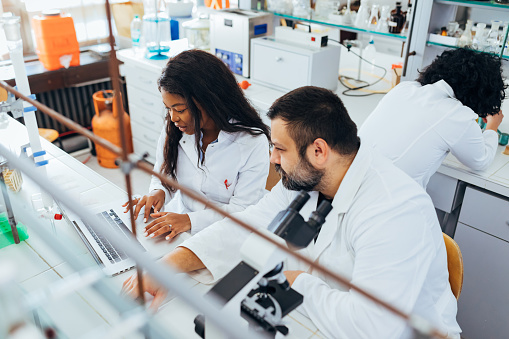 Young scientist using a laptop while conducting research in a laboratory, while her male colleague is working on the microscope