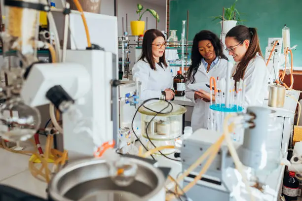 The female biochemists are standing in the laboratory and one of them is recording the results of the analyzed substances in a notebook.