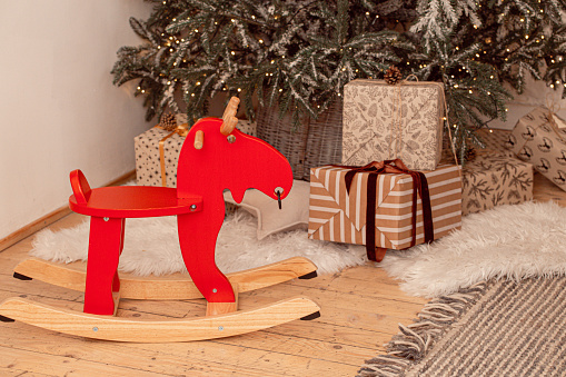 Red wooden rocking horse deer chair under Christmas tree with other wrapped presents on wooden parquet floor in modern festive interior, winter family holidays and preparing Christmas gifts concept