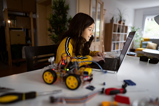 Curious and smart schoolgirl, testing her autonomous self-driven robotic car with sensors via open-source hardware and software platform on her laptop