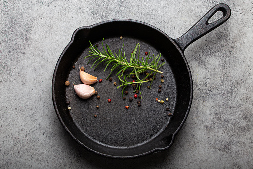 Black cast iron frying pan with ingredients for cooking: fresh green rosemary, raw garlic cloves and colorful peppercorns on gray stone background flat lay top view, cooking food concept