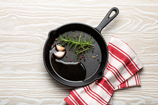 Black cast iron frying pan with ingredients for cooking: oil, fresh green rosemary, raw garlic cloves and colorful peppercorns on white rustic wooden background flat lay top view