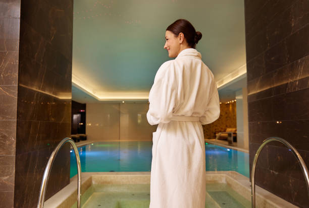 Rear view of a dark-haired beautiful woman in white terry robe standing by the pool and hot tub in luxury health spa enjoying relaxation and self-care. Beauty treatment, relaxation, body care concept Rear view of a dark-haired charming woman in white terry robe standing by the pool and hot tub in luxury health spa enjoying relaxation and self-care. Beauty treatment, relaxation, body care concept health farm stock pictures, royalty-free photos & images