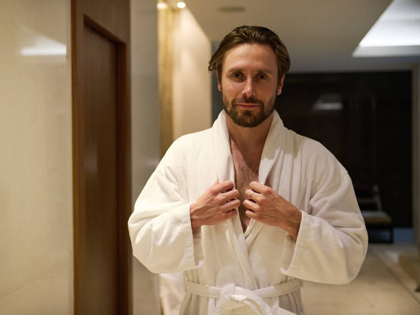 Attractive middle aged man with a charming look and a slight unshaven, handsome macho wearing white terry bathrobe, standing in a lobby of a luxury wellness spa complex, looking at camera