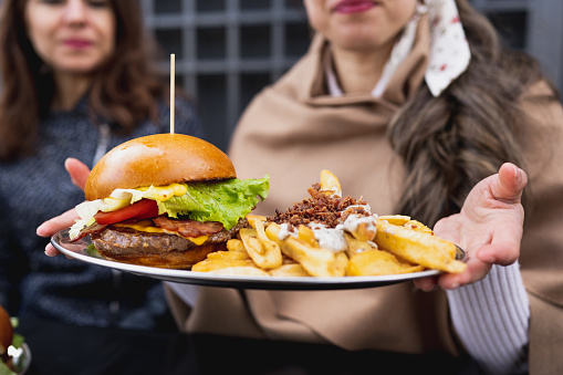 Close-up cropped woman showing plate of gourmet bacon burger with french fries and sauce. Unrecognizable females on background