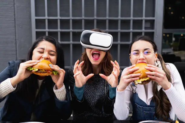 Photo of Front view shot portrait with three young women sitting at restaurant eating burgers looking at camera with virtual reality glasses.