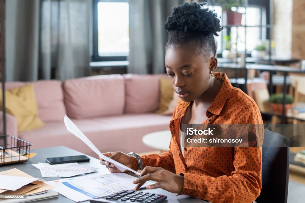 Blackwoman Calculating Tax Bill Concentrated young Blackwoman in casual shirt sitting at table in living room and calculating tax bill 20-24 Years Stock Photo