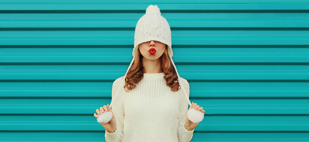 Winter portrait of beautiful young woman with pulled hat over her eyes blowing her lips sending air kiss wearing a white knitted sweater, hat on blue background