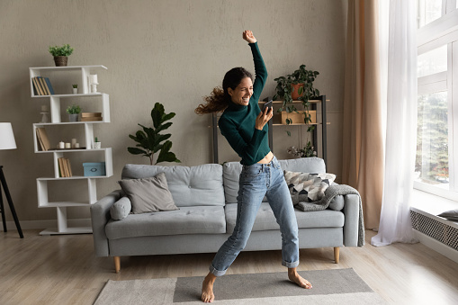 Full of energy. Active young latin lady dance at living room alone do fitness exercise by live music from cellphone. Energetic joyful millennial woman renter tenant jumping having fun at home interior