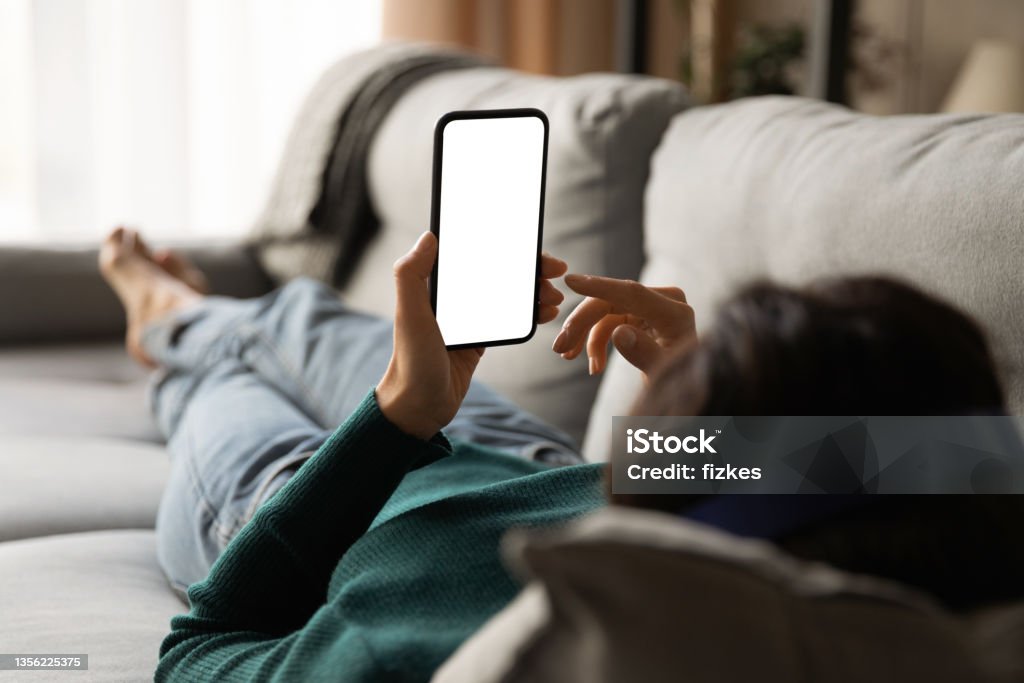 Young woman lying on sofa holding smartphone with empty screen Female using phone. Over shoulder view of young woman lying on sofa hold smartphone with blank empty screen. Template for web app chat interface online advertisement mobile game social network profile Telephone Stock Photo